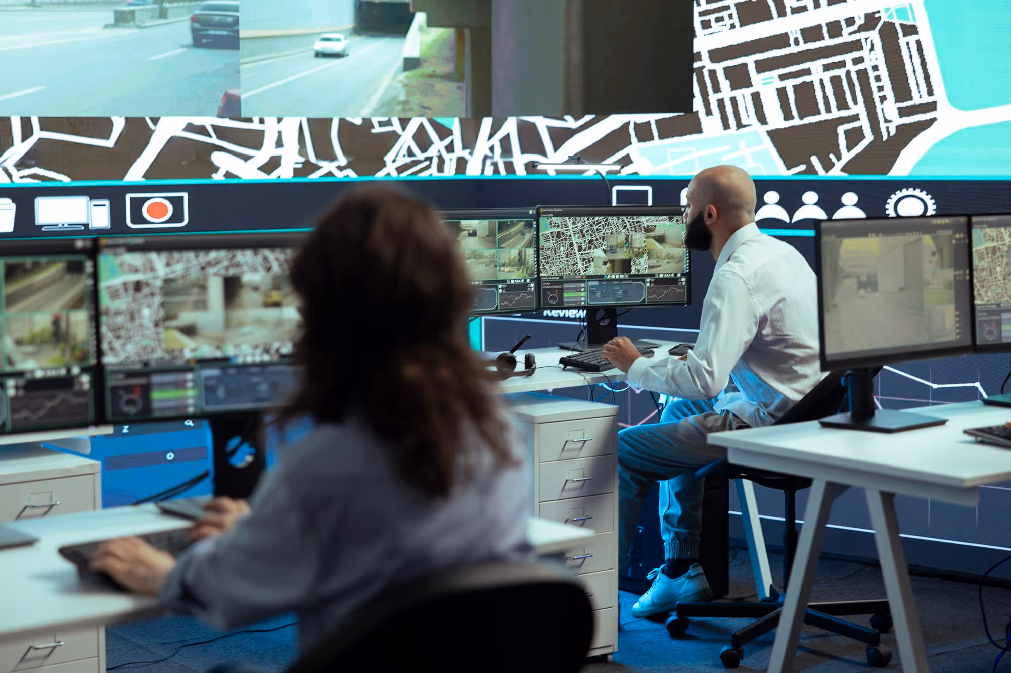 Two security operators monitoring multiple surveillance and map screens in a control room.