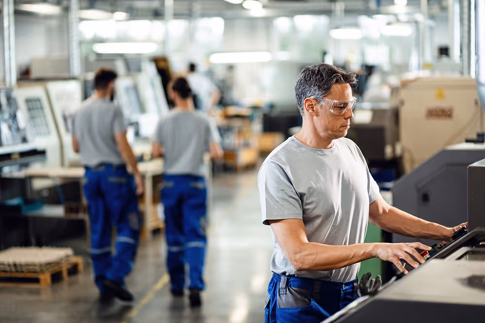Worker wearing safety glasses operating industrial machinery in a factory while two other workers walk in the background.