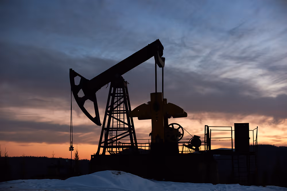 Silhouette of an oil pumpjack against a colorful sunset sky with snow on the ground.