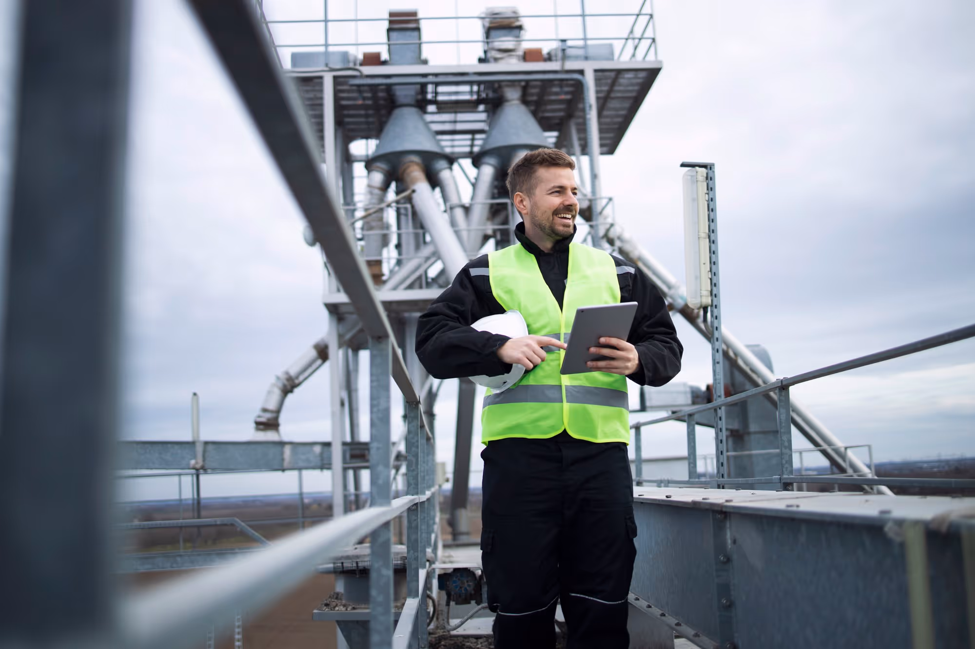 Man in a fluorescent safety vest holding a tablet and white helmet on an industrial platform with metal structures.