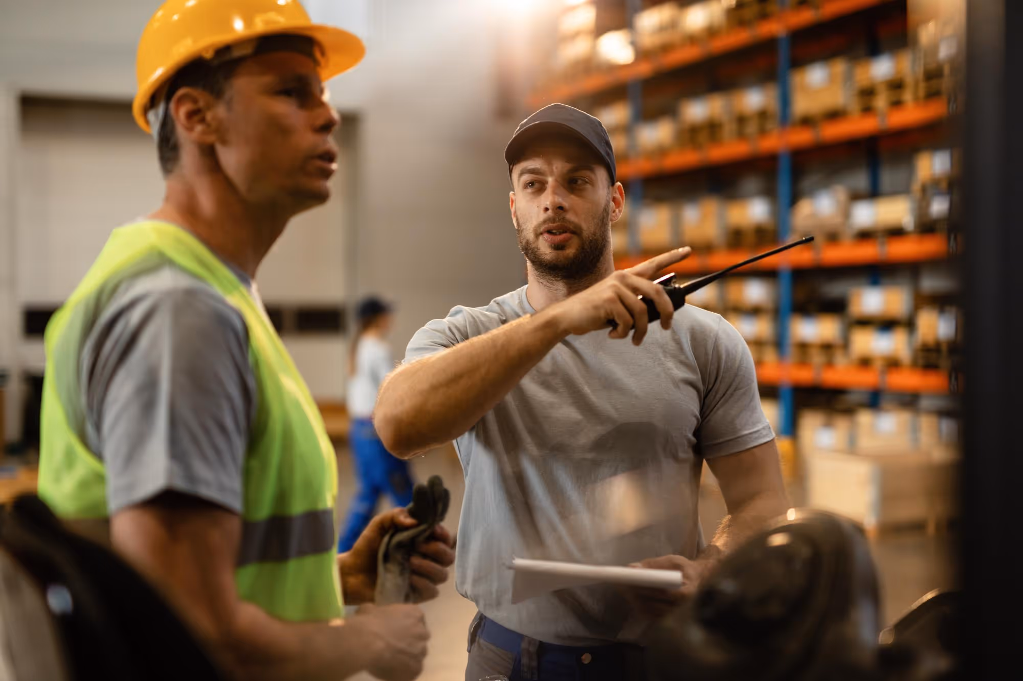 Warehouse workers discussing tasks, one pointing with a walkie-talkie and holding a clipboard.