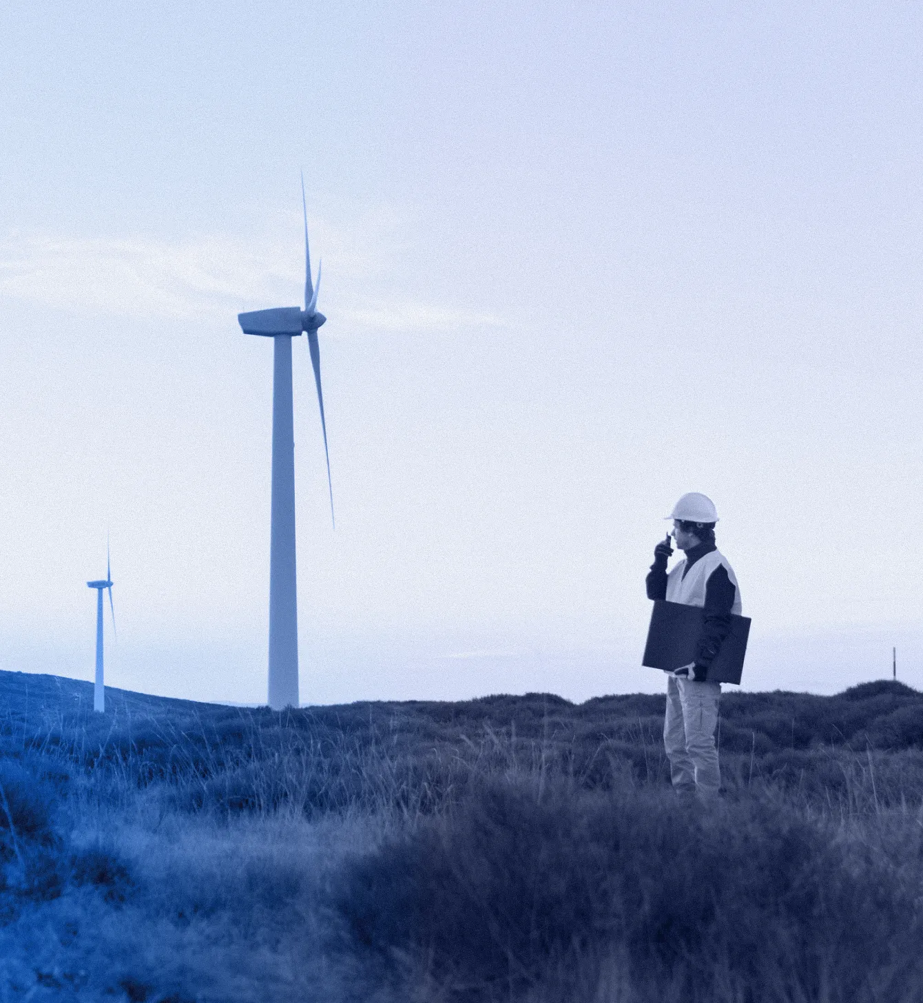 Engineer in a safety helmet and vest using a walkie-talkie near wind turbines on a grassy hill.