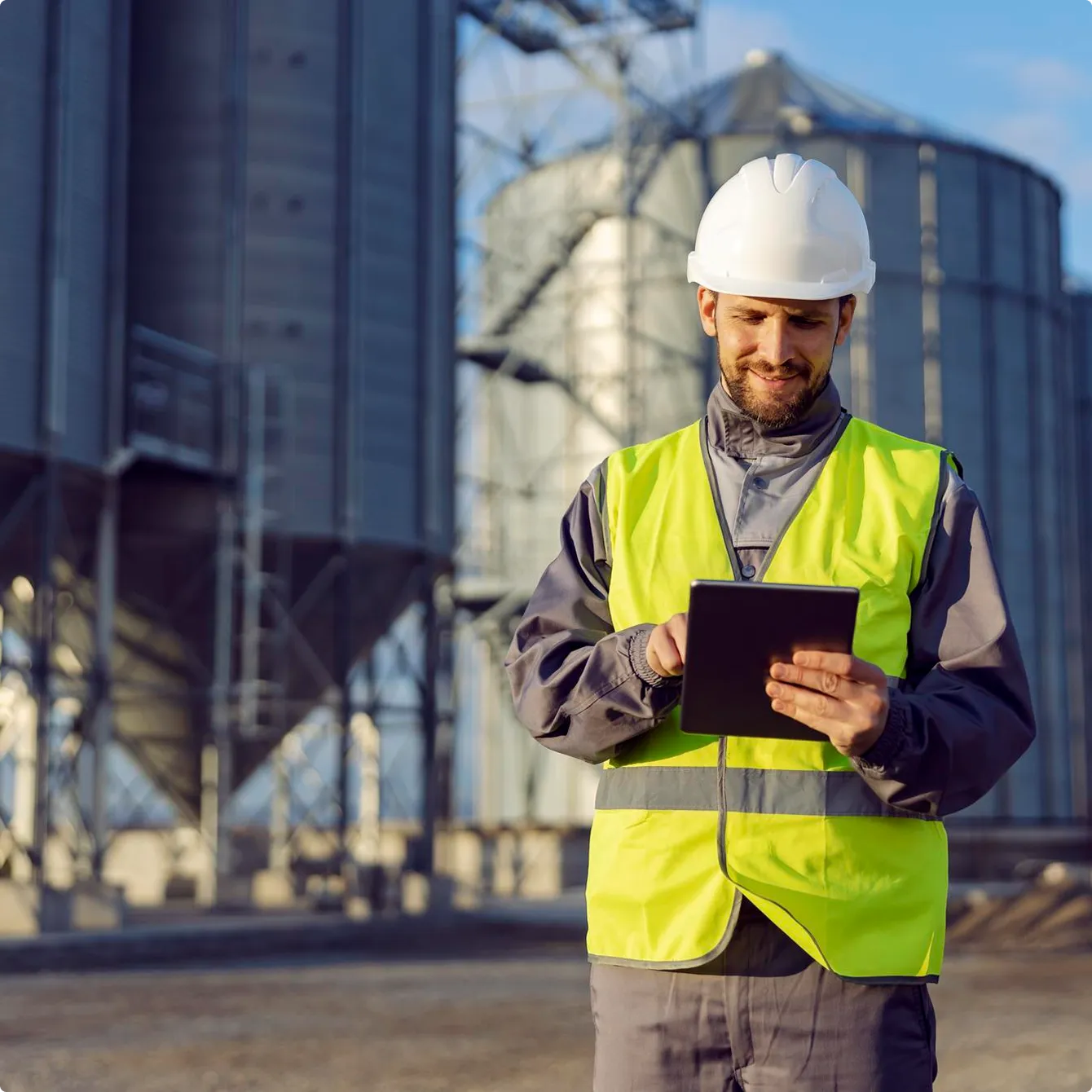 Construction worker in a white hard hat and yellow safety vest using a tablet at an industrial facility with large metal silos.