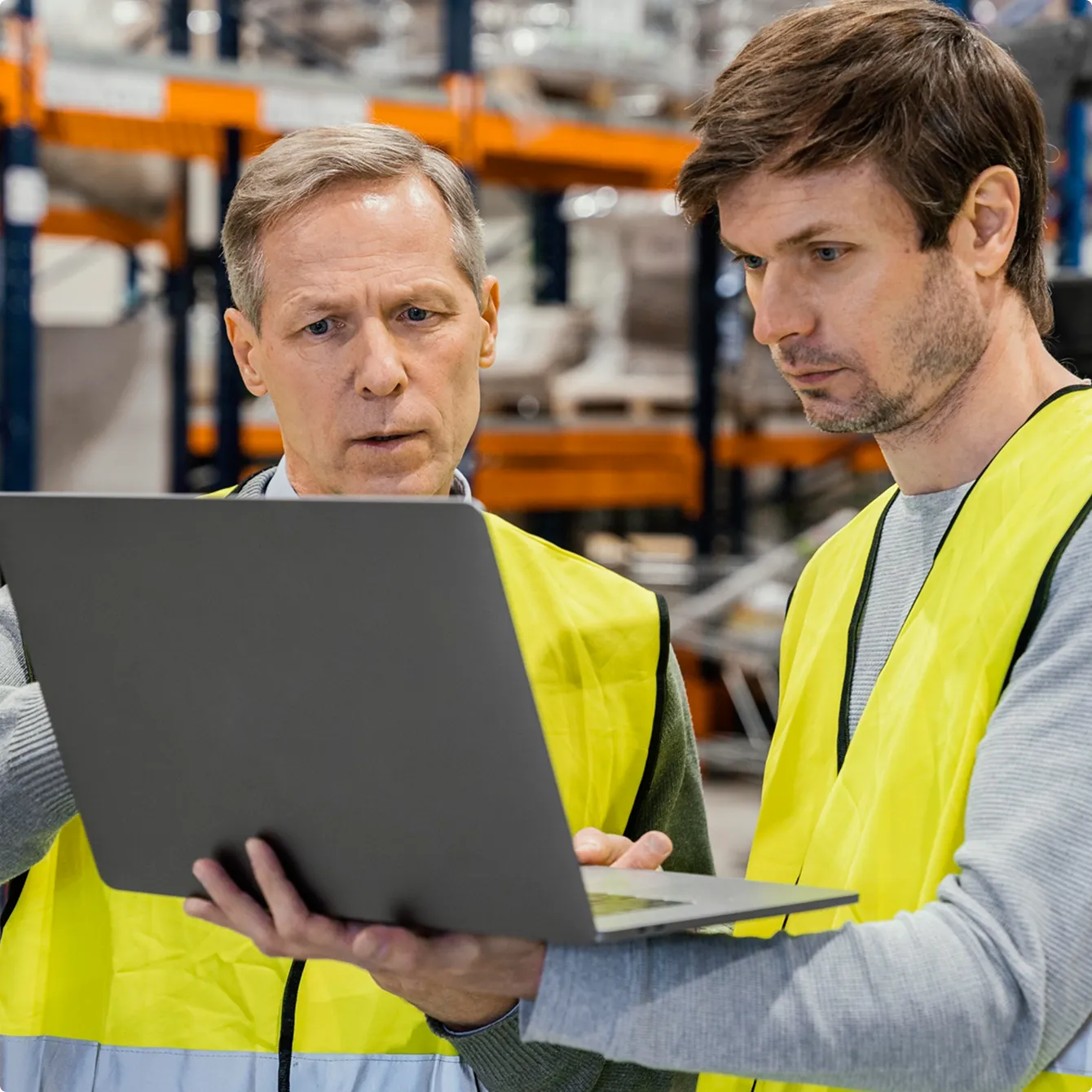 Two men in yellow safety vests looking at a laptop in a warehouse setting.