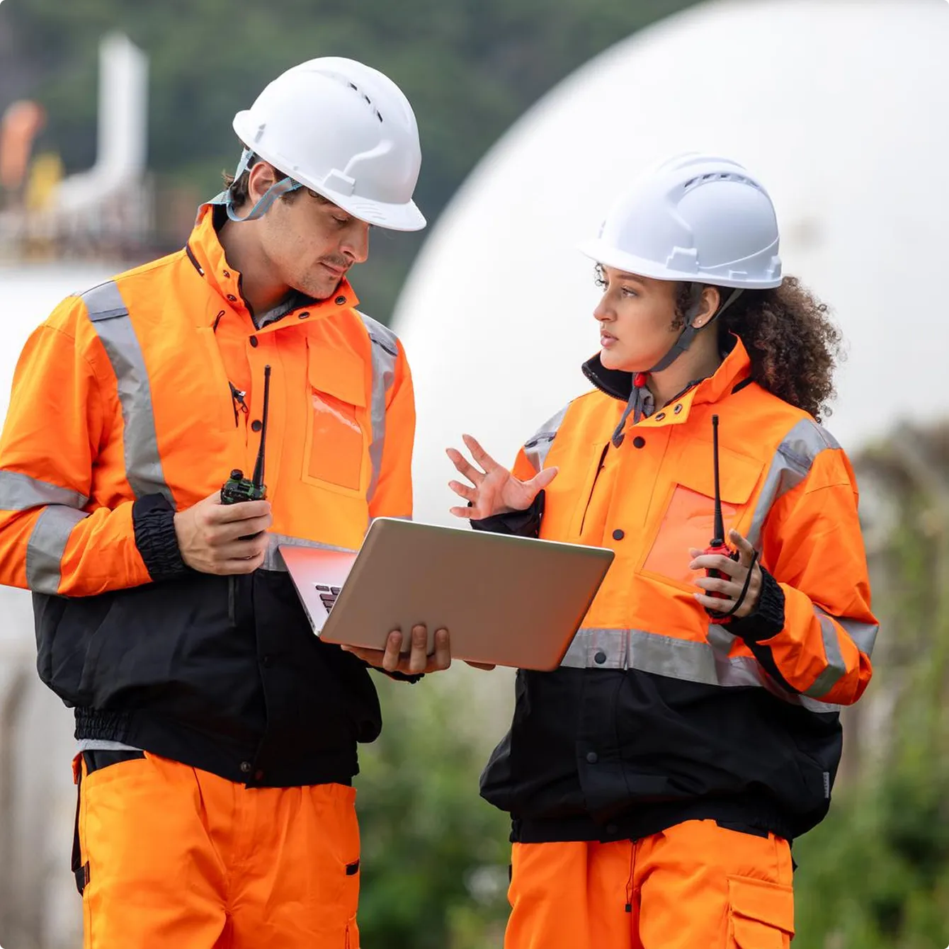 Two construction workers in orange safety gear and white helmets discussing while holding a laptop and walkie-talkies outdoors.