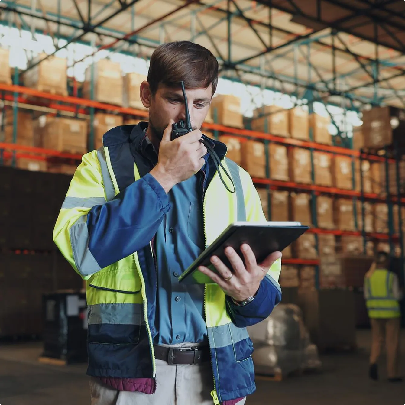 Warehouse worker in a high-visibility jacket using a walkie-talkie and holding a tablet in a storage facility.