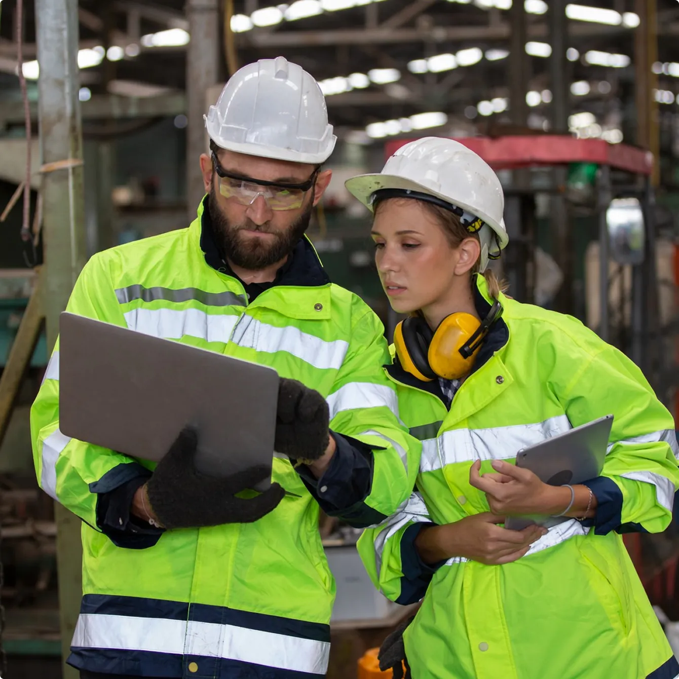 Two factory workers in safety gear, including white helmets and neon yellow jackets, reviewing information on a laptop and tablet inside an industrial setting.