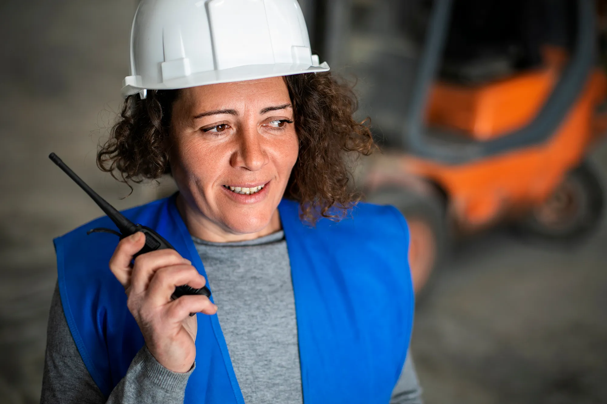 Construction worker wearing a white hard hat and blue vest talking on a walkie-talkie with blurred orange machinery in the background.