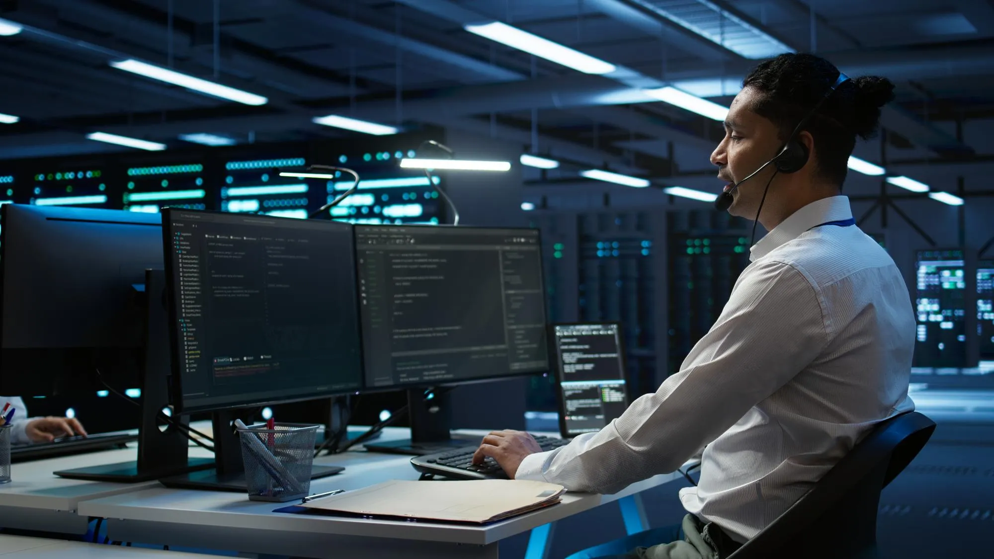 Man in a white shirt with a headset working at a desk with multiple monitors displaying code in a dimly lit server room.