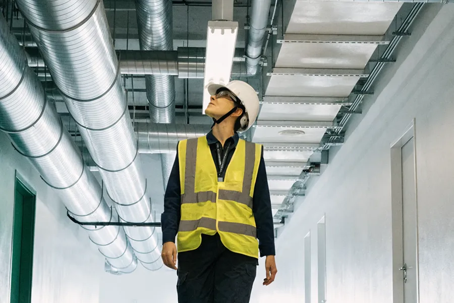 Construction worker wearing a white hard hat and yellow safety vest inspecting overhead HVAC ducts in an industrial hallway.