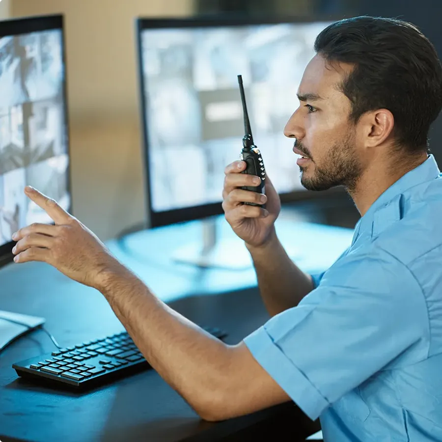 Man in blue shirt using a walkie-talkie while pointing at one of two computer monitors displaying surveillance footage.