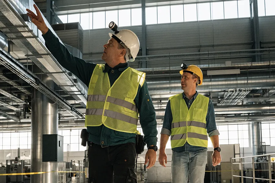 Two industrial workers wearing hard hats, headlamps, and reflective vests inside a factory, with one pointing upward.