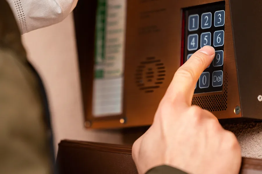 Person wearing a mask pressing number six on a security keypad intercom mounted on a wall.