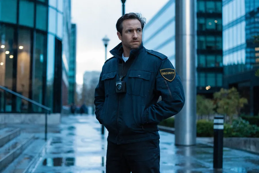 Security guard in black uniform and cap standing at a gated entrance holding a walkie-talkie.