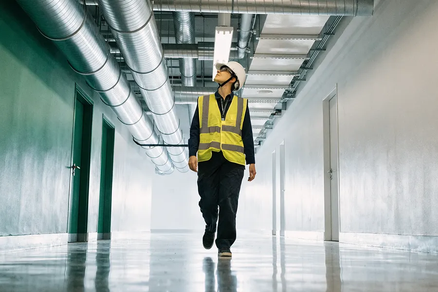 Construction worker wearing a helmet and safety vest walking in a hallway with exposed ceiling ducts.