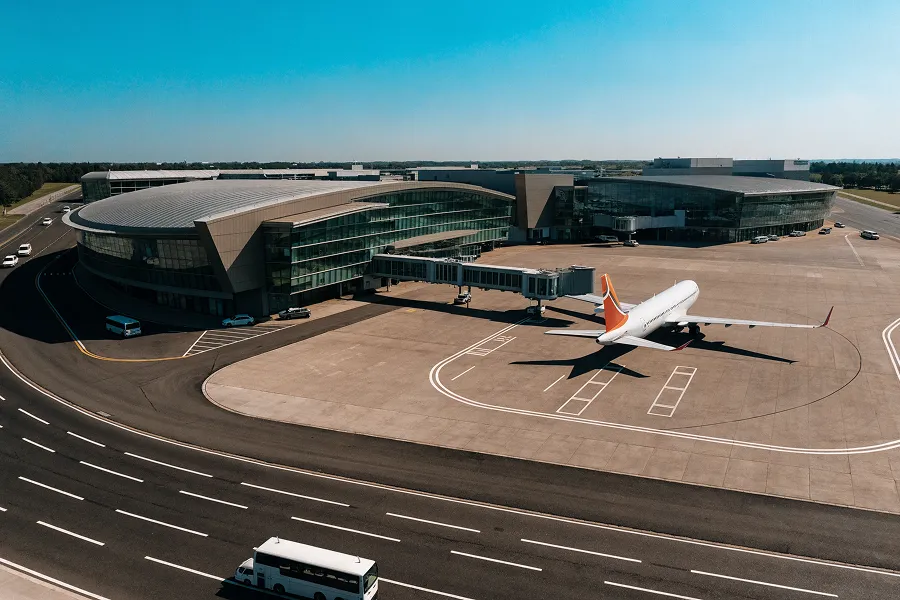 A modern airport terminal with a jet bridge connected to a parked airplane on the tarmac under clear blue sky.
