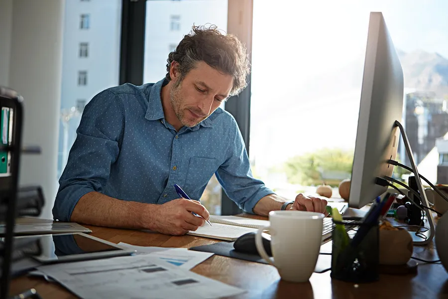 Man with curly hair in blue shirt writing in a notebook at a desk with a computer and papers.