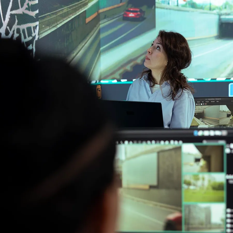 Woman with dark hair looks up while standing in front of large computer screens showing road and traffic images.