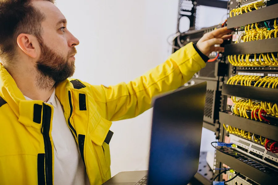Man in a yellow jacket working on a server rack with network cables, using a laptop.