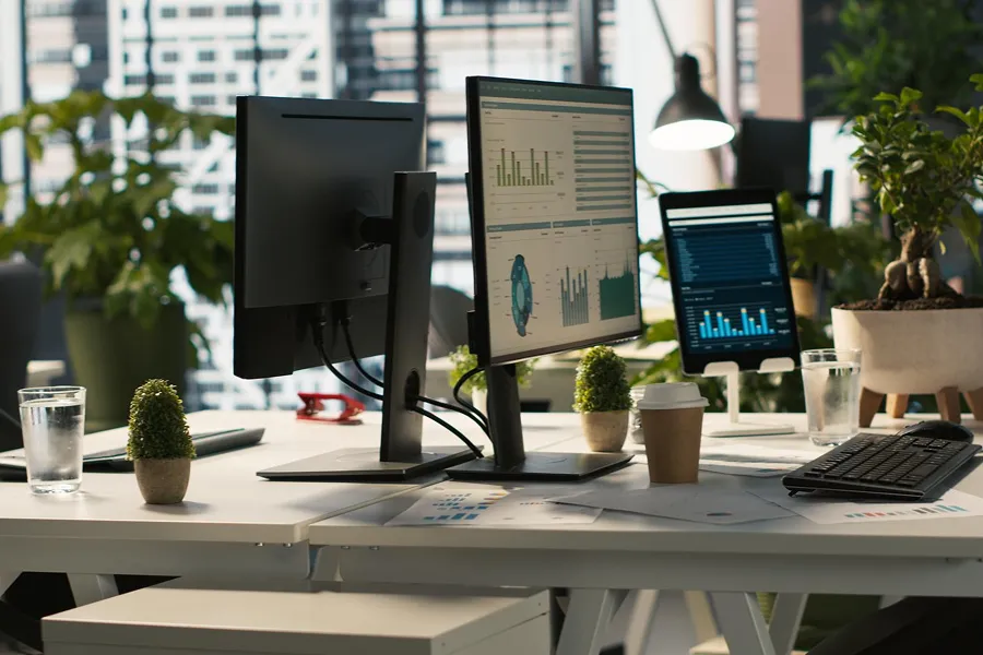 Modern office desk with dual monitors, a tablet displaying charts, plants, and work documents in a bright workspace.