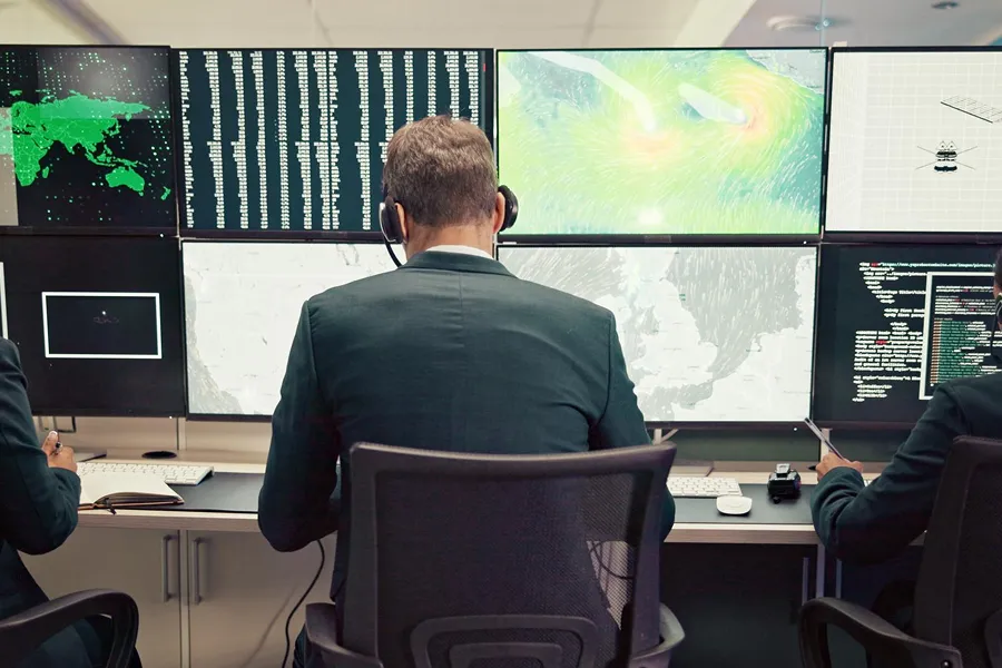 Three professionals wearing headsets sitting in front of multiple monitors displaying maps, data, and code in a control room.