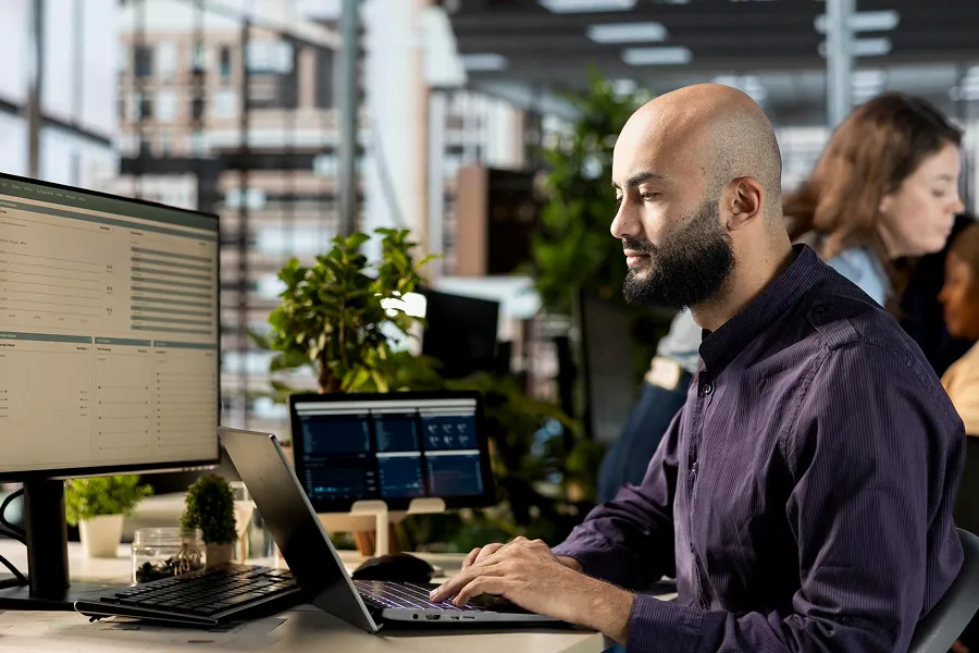 Bearded man working on a laptop at a desk with dual monitors in a modern office.