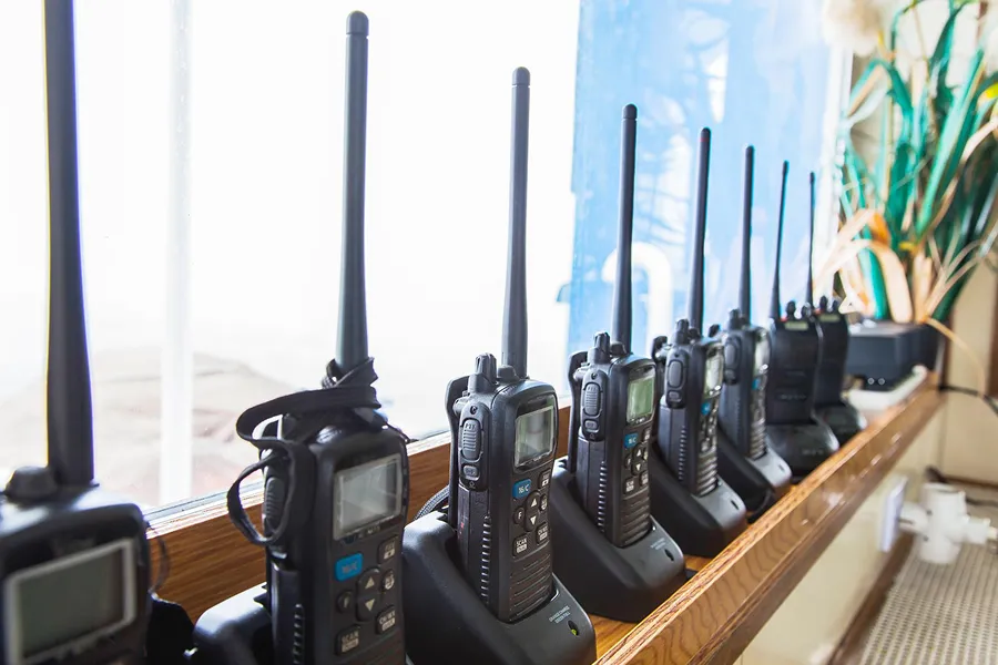 Row of black walkie-talkies with antennas standing on charging docks near a window.