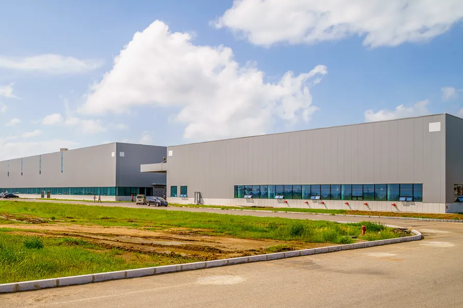 Large modern industrial warehouse building under a partly cloudy sky with a paved road and green grass in the foreground.