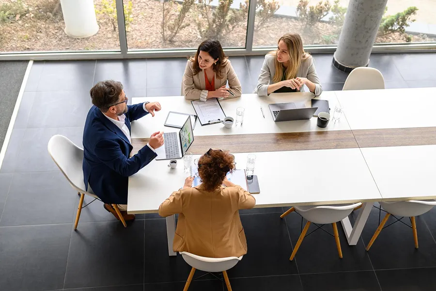Four business professionals having a meeting around a white table with laptops, tablets, and documents in a modern office with large windows.