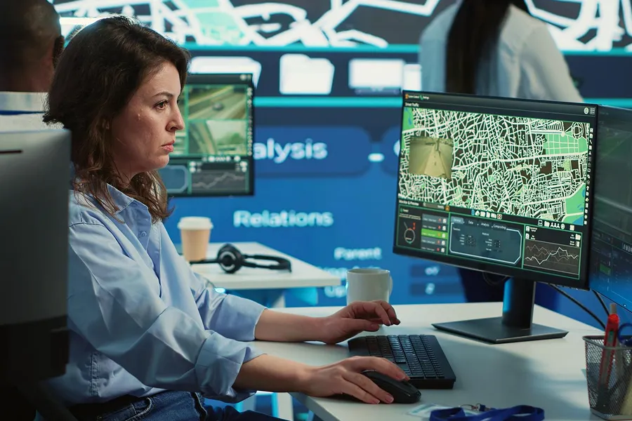Woman analyzing a detailed digital map on a computer screen in a control room with other people and technology in the background.