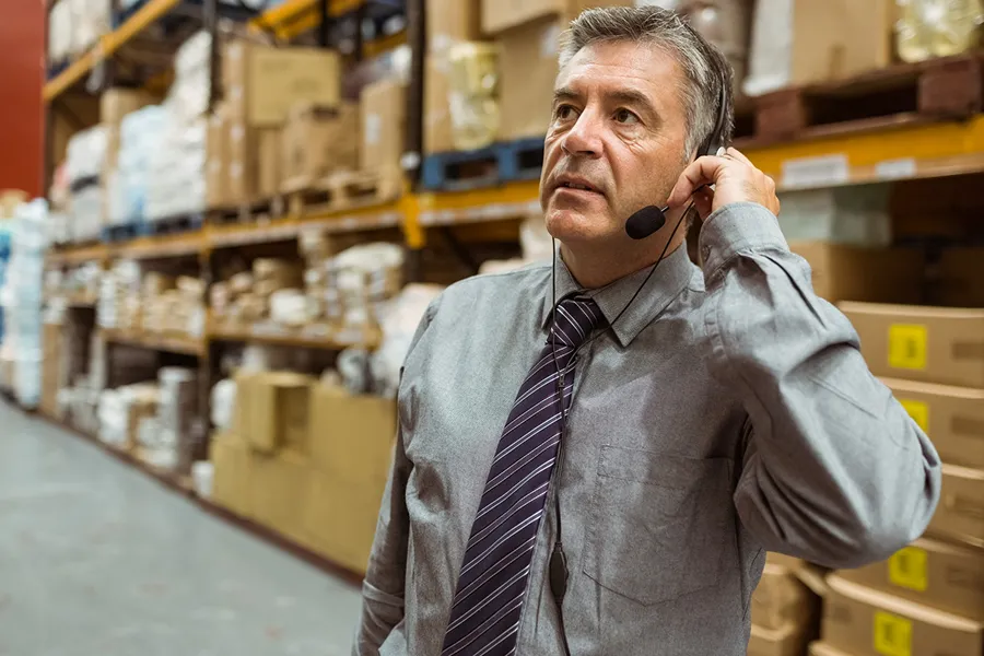 Man wearing headset with microphone standing in a warehouse with shelves of boxes and packages.