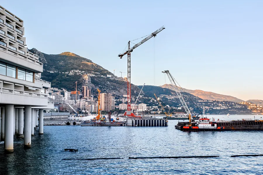Coastal construction site with cranes near the water, buildings, and mountain in the background during clear daytime.