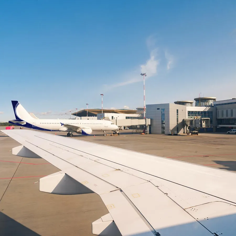 View of an airport runway and terminal with a parked airplane, seen from inside another plane showing its wing.