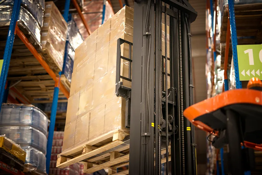 Forklift lifting a pallet wrapped in plastic in a warehouse with high shelves stacked with goods.