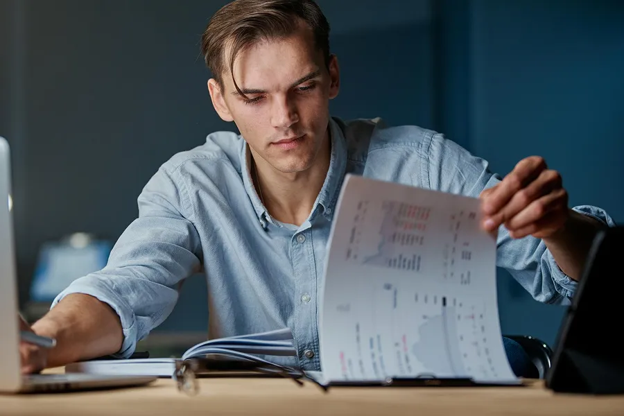 Young man in a blue shirt analyzing financial charts and documents at a desk with a laptop.