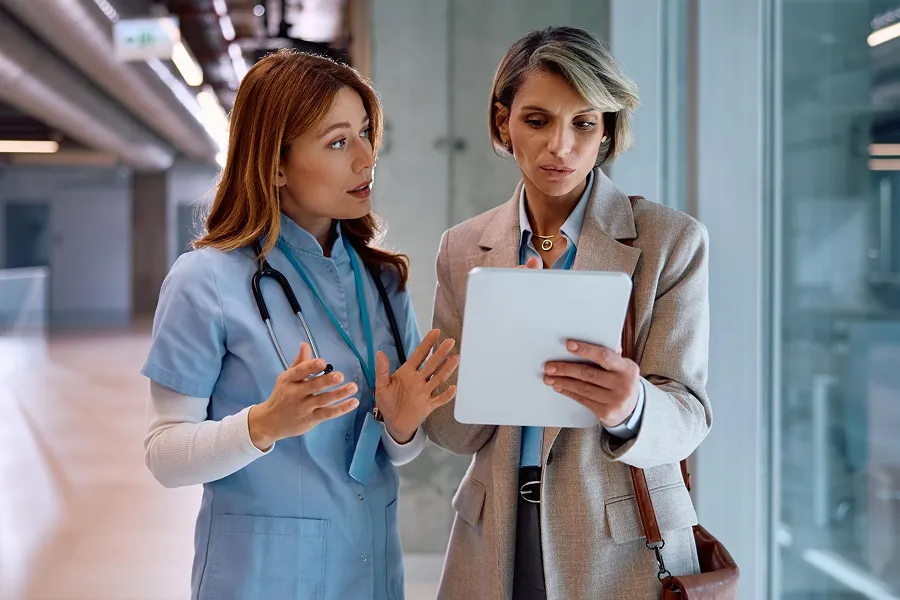 Female healthcare professional in blue scrubs with a stethoscope discussing information on a digital tablet with a woman in business attire in a hospital corridor.