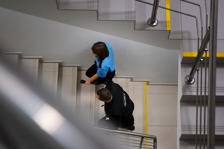 Top view of a security guard escorting a woman down a stairwell with metal railings and yellow safety strips on steps.