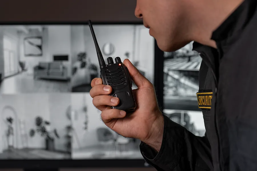 Security guard holding a walkie-talkie with multiple surveillance camera feeds displayed on a monitor in the background.