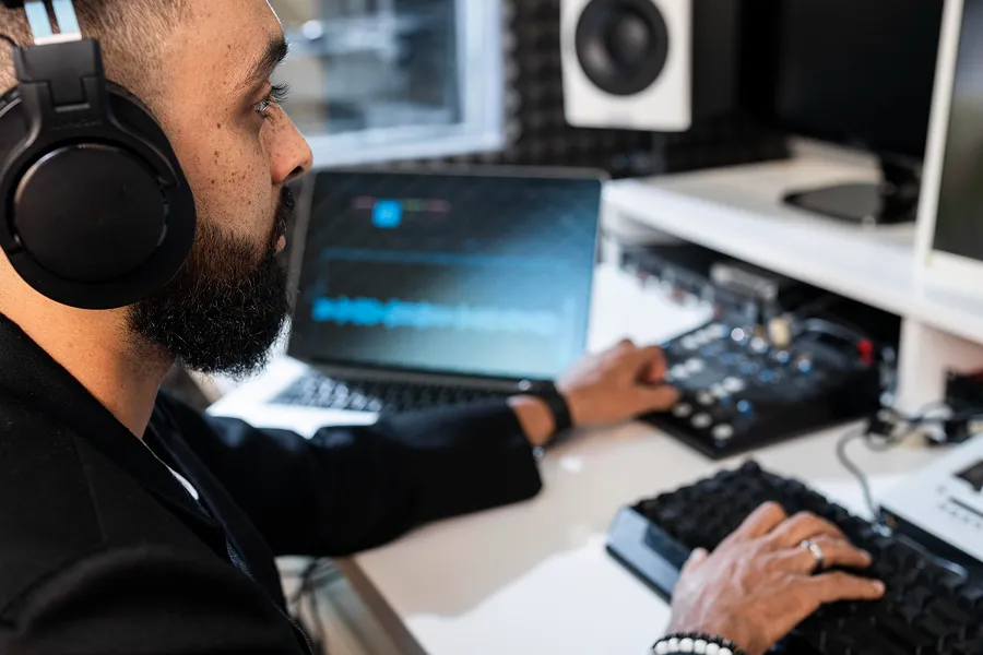 Man wearing headphones working on audio editing using a laptop and sound mixer in a studio.