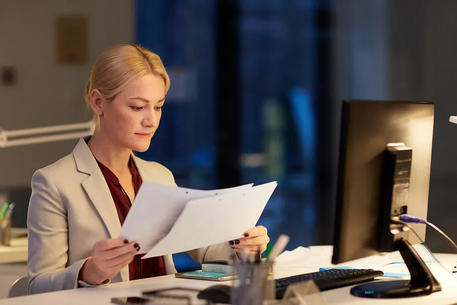 Woman with blonde hair wearing a beige blazer and maroon shirt reviewing documents at an office desk with a computer monitor.