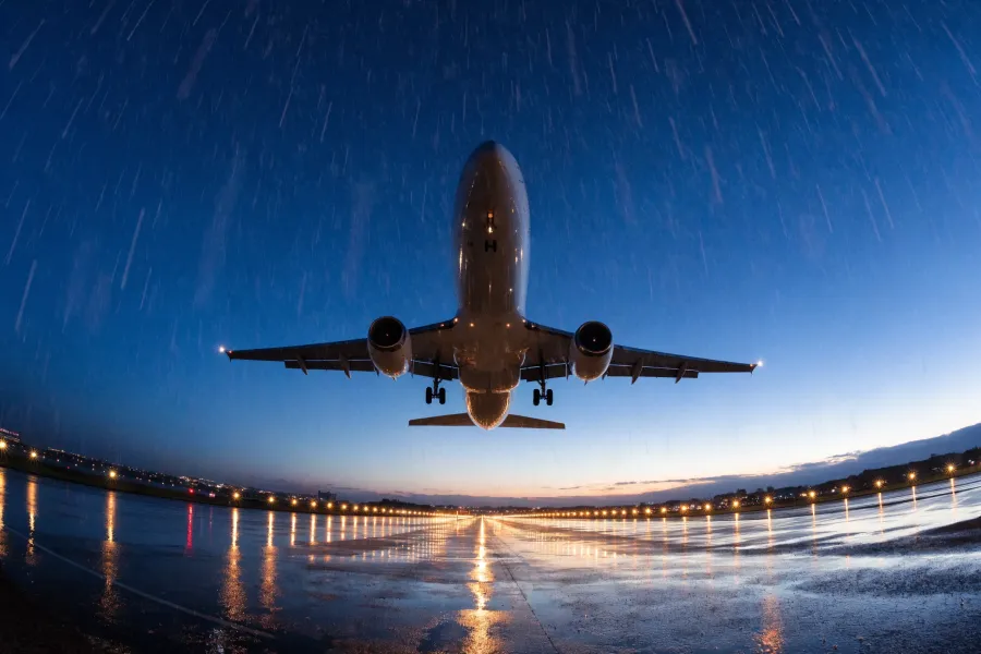 Airplane landing on wet runway at dusk with rain falling and lights reflecting on the surface.