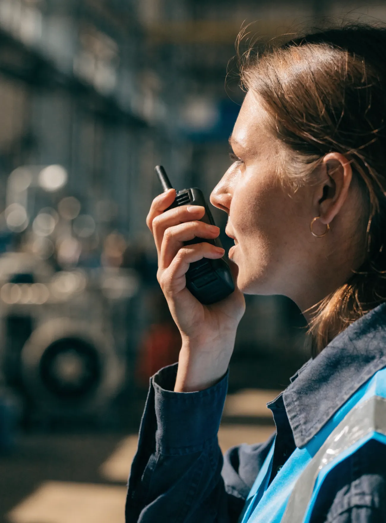 Close-up of a woman in workwear speaking into a walkie-talkie in an industrial setting.