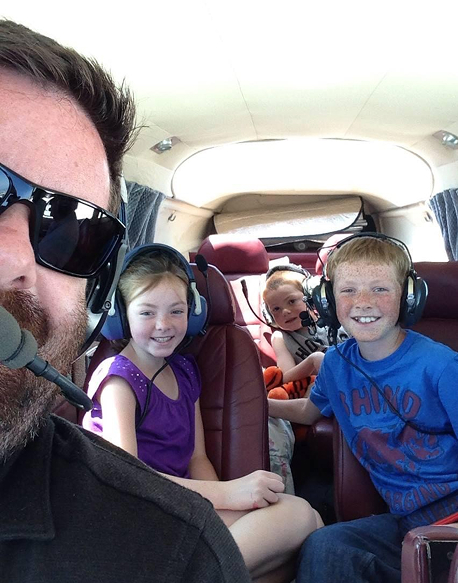 Man wearing sunglasses and headset taking a selfie with three smiling children seated inside an aircraft wearing headsets.