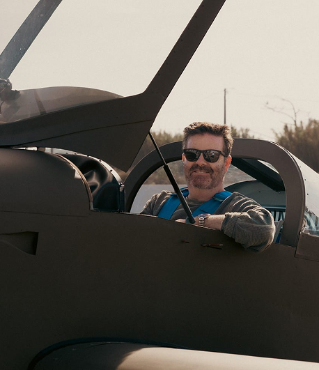 Man wearing sunglasses and a blue harness sitting inside a vintage aircraft cockpit with the canopy open.