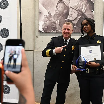 A male police officer in uniform pointing and smiling next to a female police officer holding an award and certificate, while being photographed.