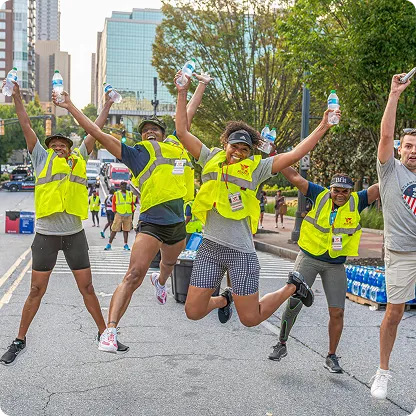 Group of smiling volunteers in yellow vests jumping and holding water bottles on a city street during an event.