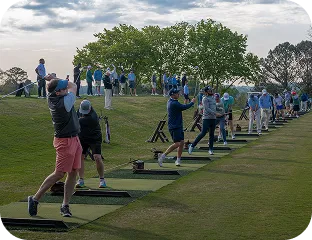 Golfers practicing swings on a driving range with multiple hitting stations lined up under trees.