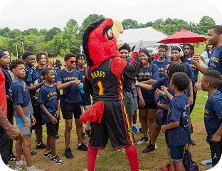 Children wearing matching navy blue shirts gathered outdoors around a red bird mascot in a black sports jersey numbered 1.