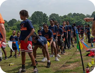 Group of children in navy blue shirts standing in a line outdoors on grass near colorful flags.