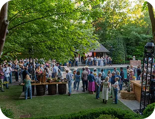 Outdoor gathering of people socializing near a pool with trees and a pavilion in the background during daylight.
